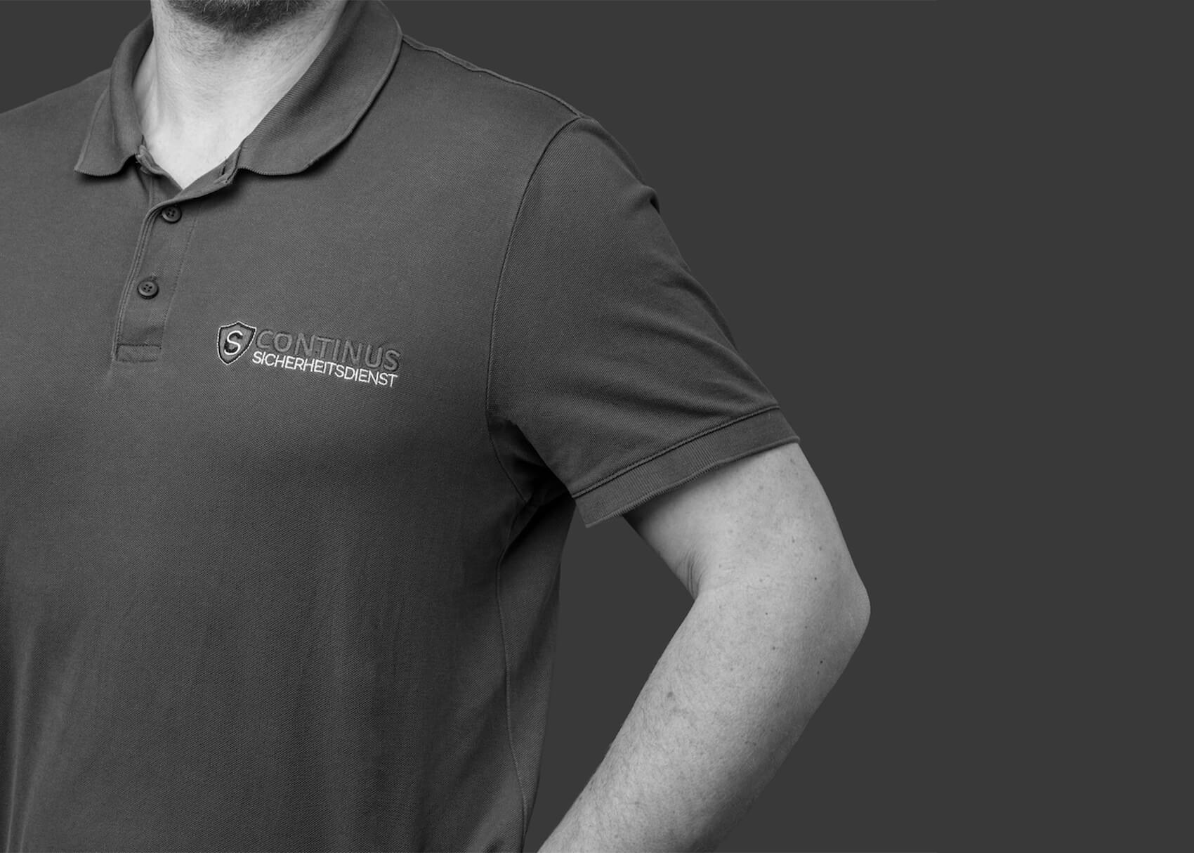 Schwarz-weiß Close-up eines dunkelgrauen Shirts von Continus Sicherheitsdienst mit klar sichtbarem Logo, minimalistisches Corporate Design.