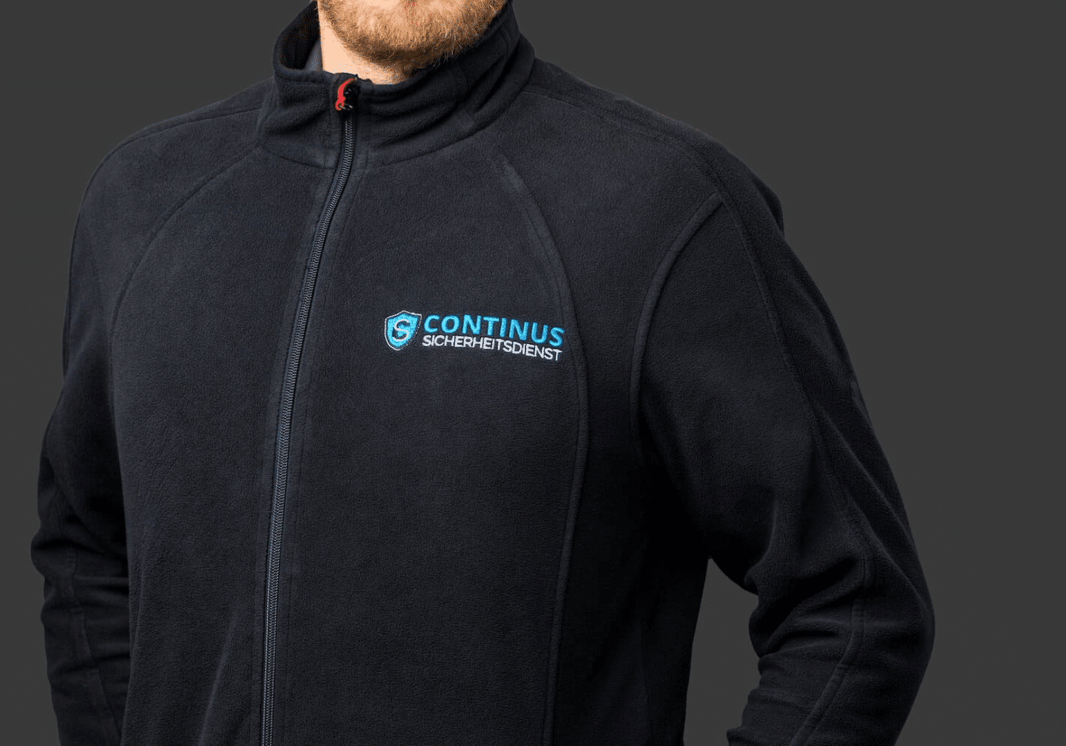 Oberkörperausschnitt eines Mannes mit Bart der eine dunkelblaue Fleecejacke mit dem Continus-Sicherheitsdienst-Logo trägt.