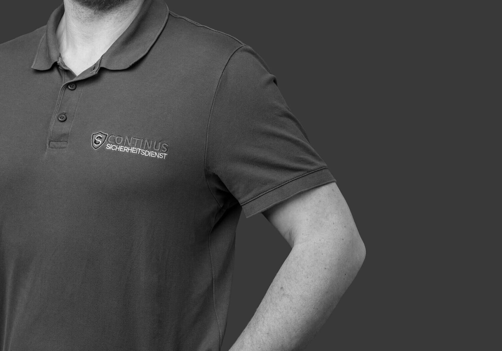 Schwarz-weiß Close-up eines dunkelgrauen Shirts von Continus Sicherheitsdienst mit klar sichtbarem Logo, minimalistisches Corporate Design.